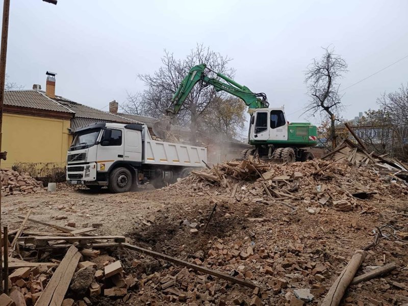 Колісний екскаватор розбирає будівлю, поруч самоскид Volvo під завантаження