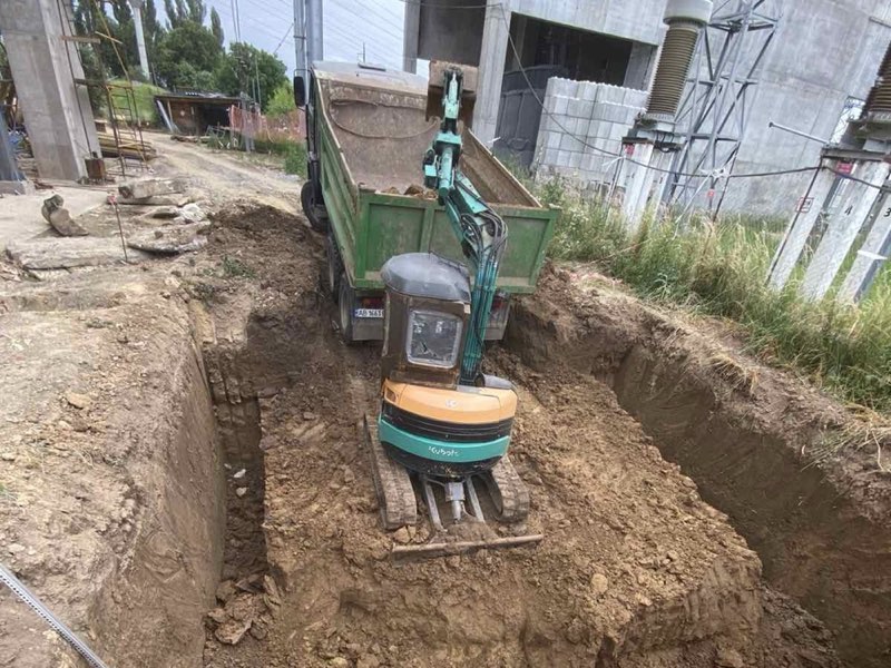 Мініекскаватор Kubota у глибокій траншеї, позаду самоскид